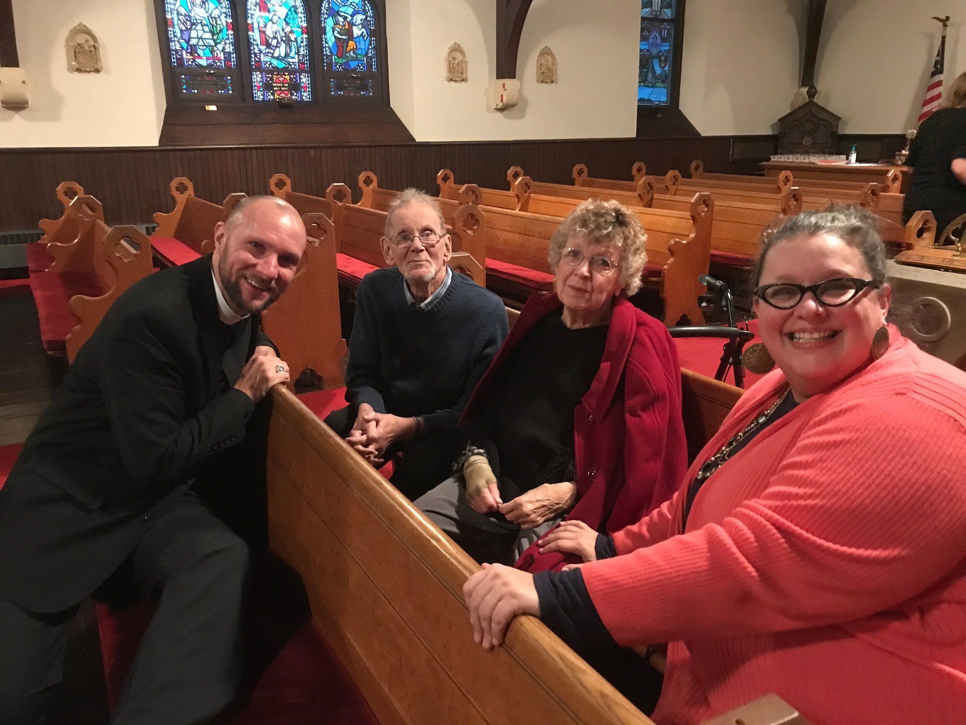 Four people smiling in church pews. The man on the left wears a black clerical collar. The woman on the right has glasses and a red sweater.