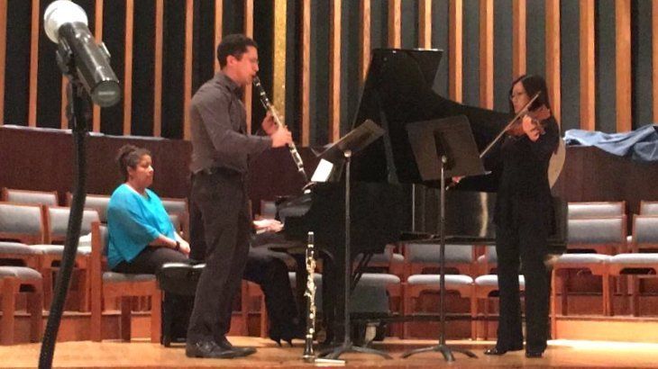 A musician playing clarinet, another playing violin, and a pianist at a performance on stage.