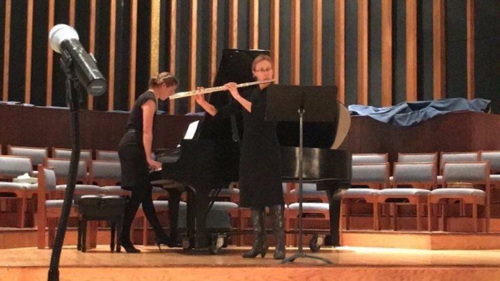 Woman playing a flute with a pianist in a concert hall. Both are dressed in black.