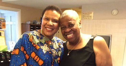 Two smiling women posing closely together indoors; one in patterned blue, the other in black.