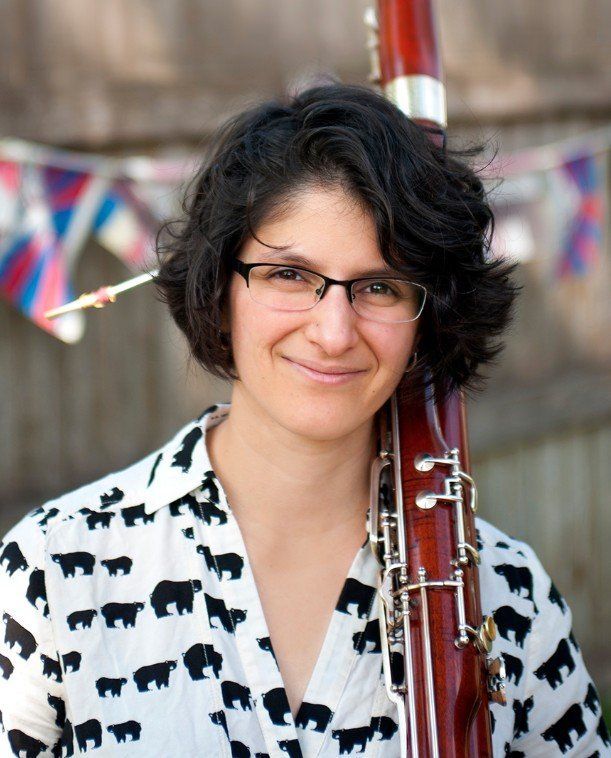 Woman with glasses holding a bassoon, smiling. Wearing a bear-patterned shirt, standing outside with flags in the background.