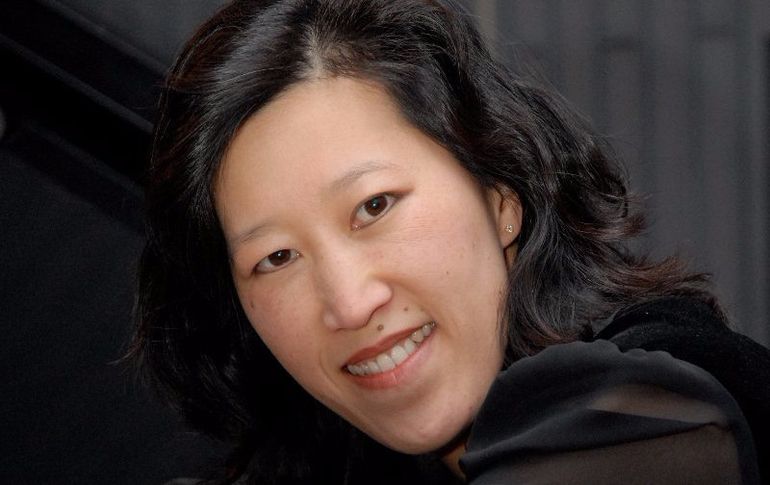 Woman with dark hair smiles, wearing dark clothing; soft focus background.