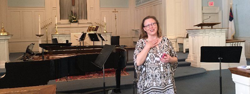 Woman in glasses speaks in a church, hand over heart, near a piano.