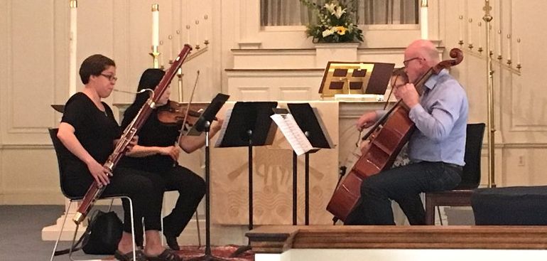 Musicians playing bassoon, violin, and cello in a light-filled church, performing together.