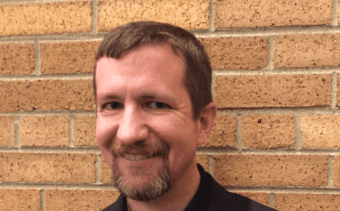 Man with short brown hair and beard smiles in front of a brick wall.
