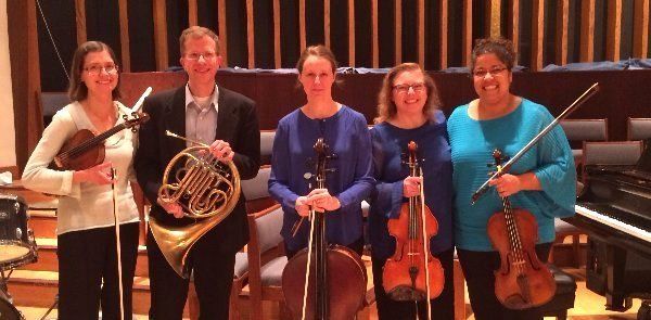 Five musicians pose with their instruments in a concert hall.