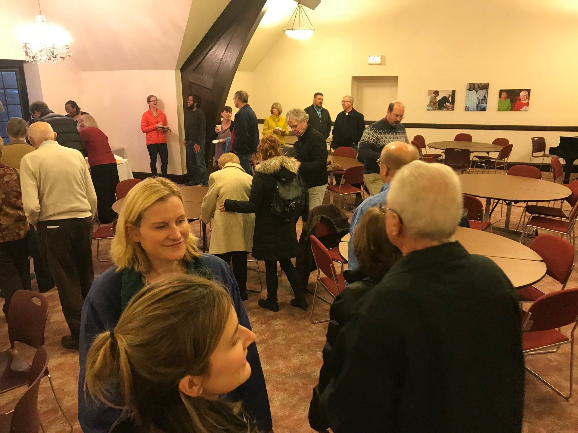 People gathered in a room with tables and a piano. Several are talking, some are wearing coats.