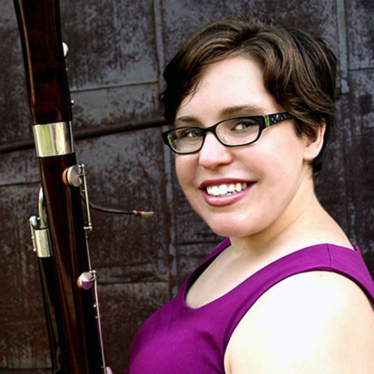 Woman with glasses smiles, holding a bassoon. She wears a purple top, set against a metal backdrop.