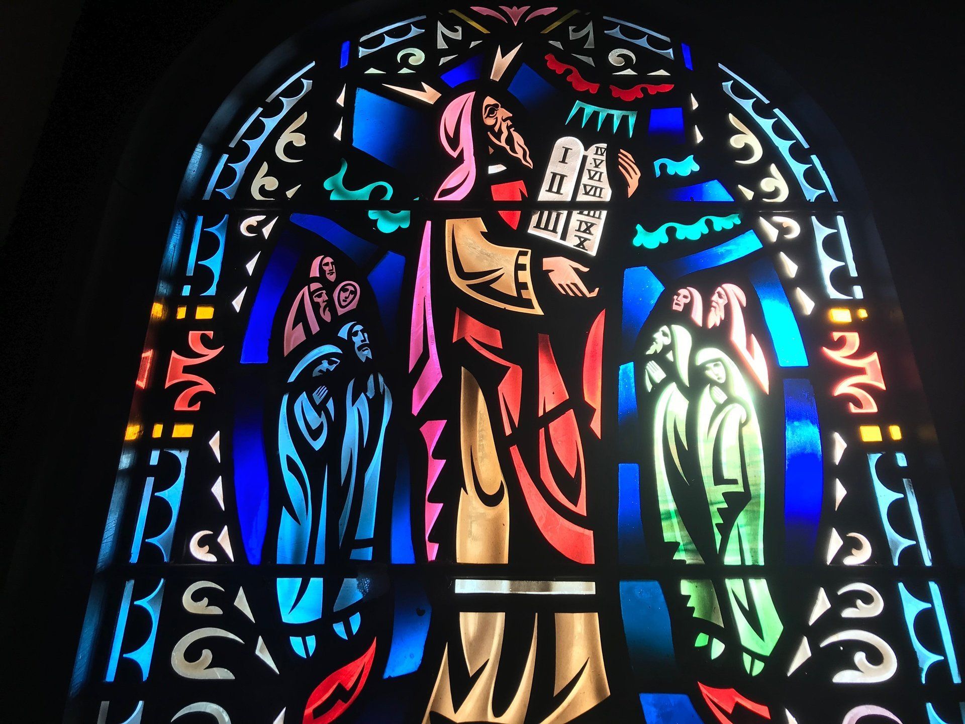 Stained glass window depicting Moses holding tablets with the Ten Commandments, flanked by figures, in vibrant colors.