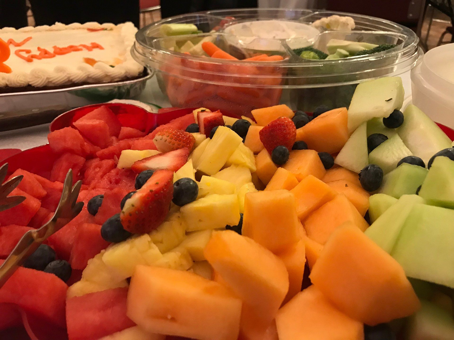 Fruit platter with watermelon, pineapple, and cantaloupe; veggie tray; cake.