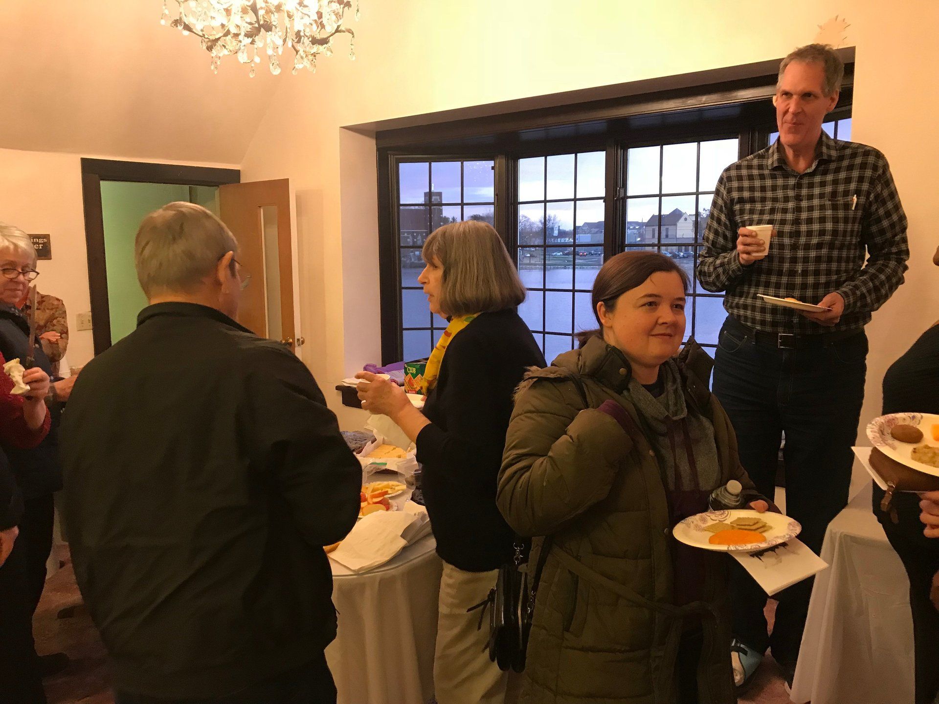 People at a gathering inside a room, some eating snacks. Windows in the background overlook a body of water.