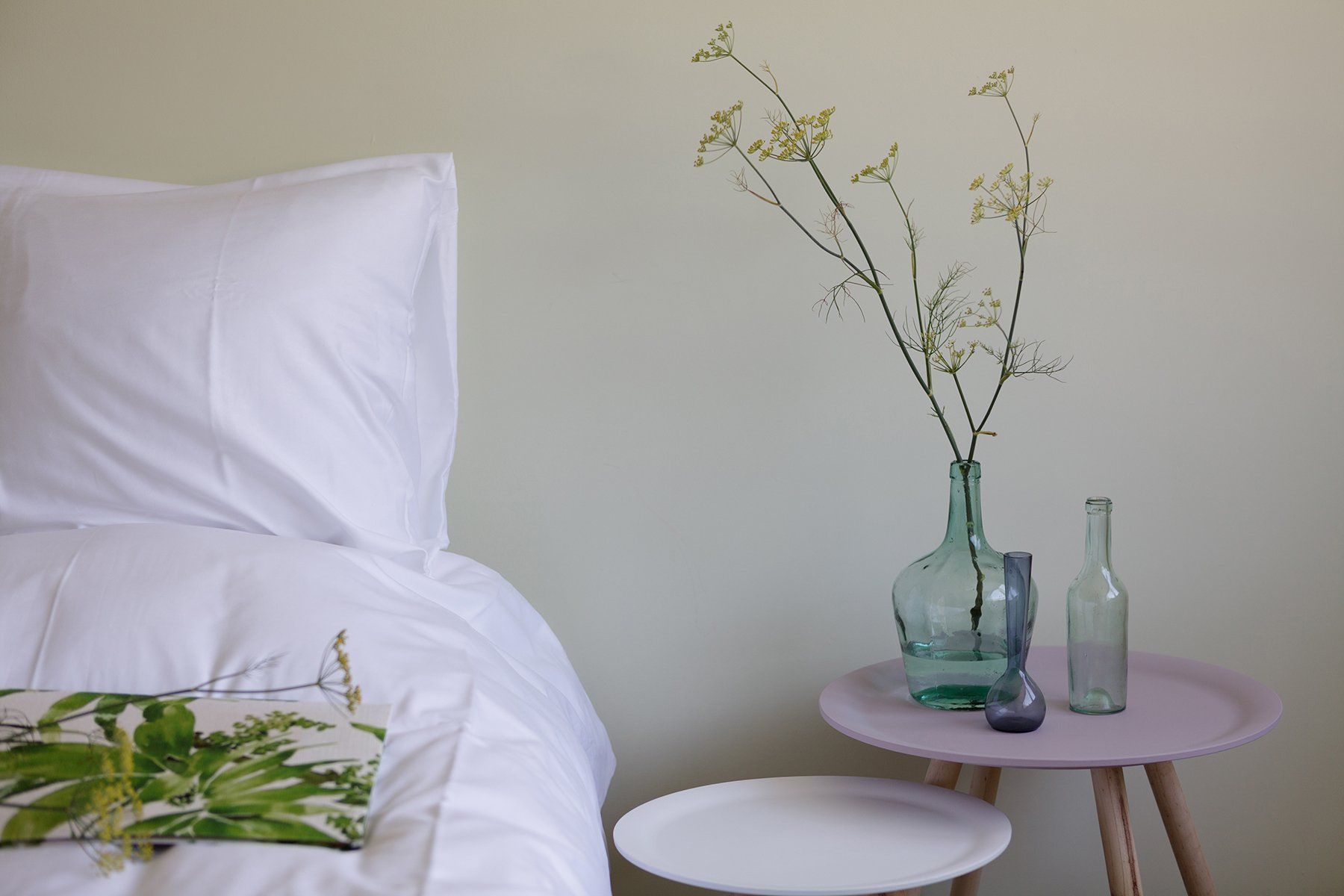 Wit beddengoed met decoratief kussen, bloemstuk op roze tafel, witte muur.