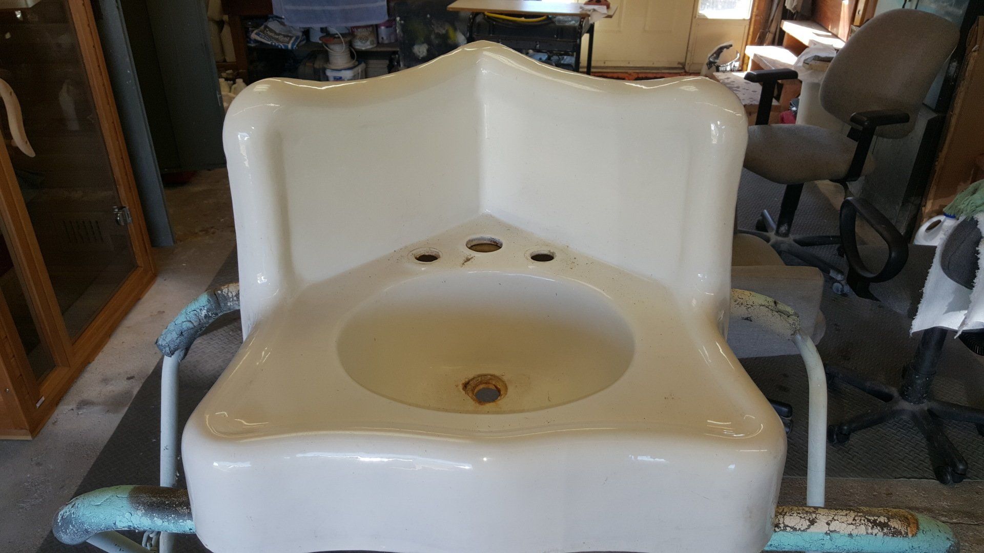 White, corner-shaped porcelain sink with three faucet holes, set on a distressed metal stand, in a cluttered indoor setting.