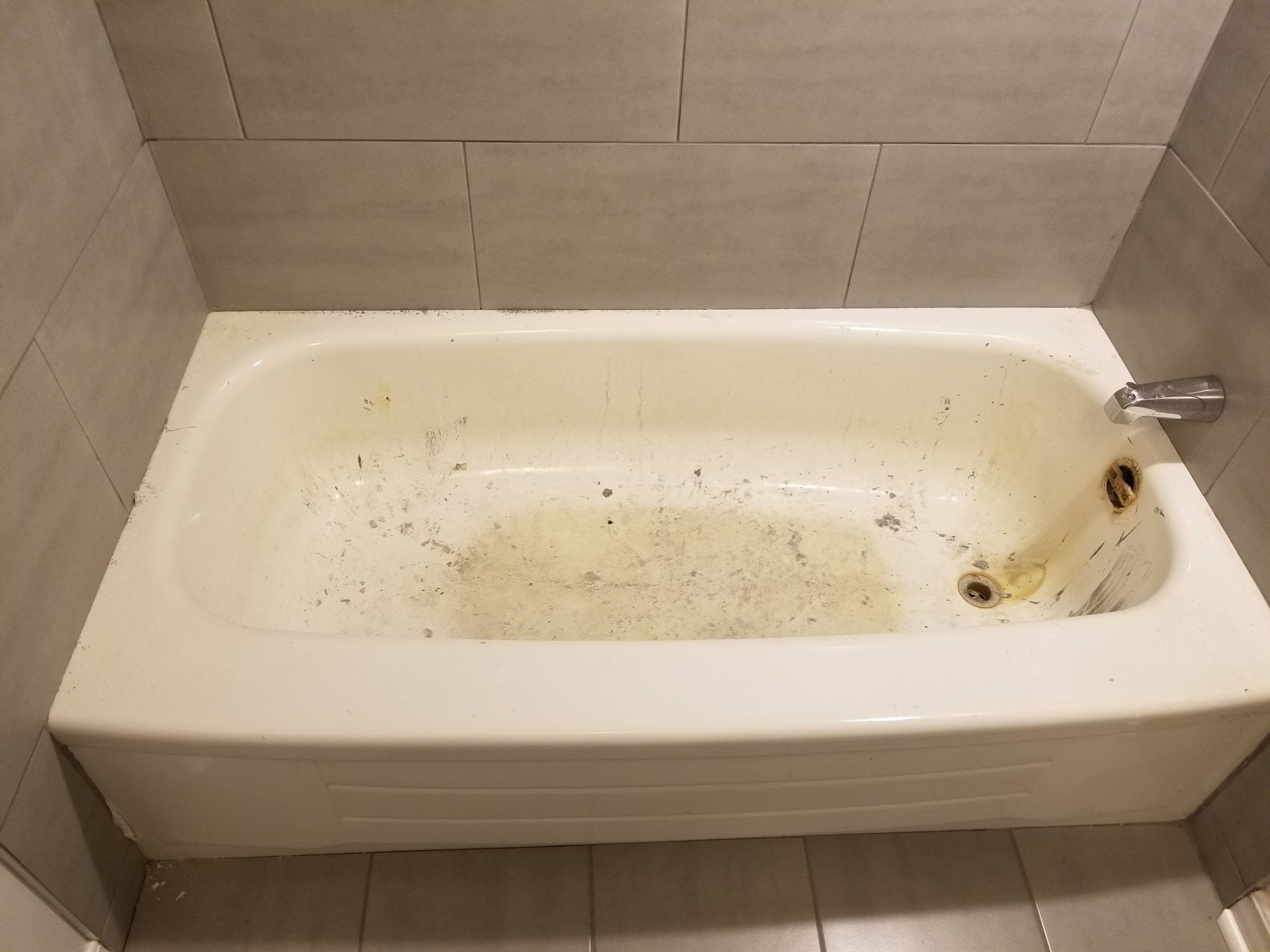 Dirty bathtub with debris in a bathroom with gray tiles.