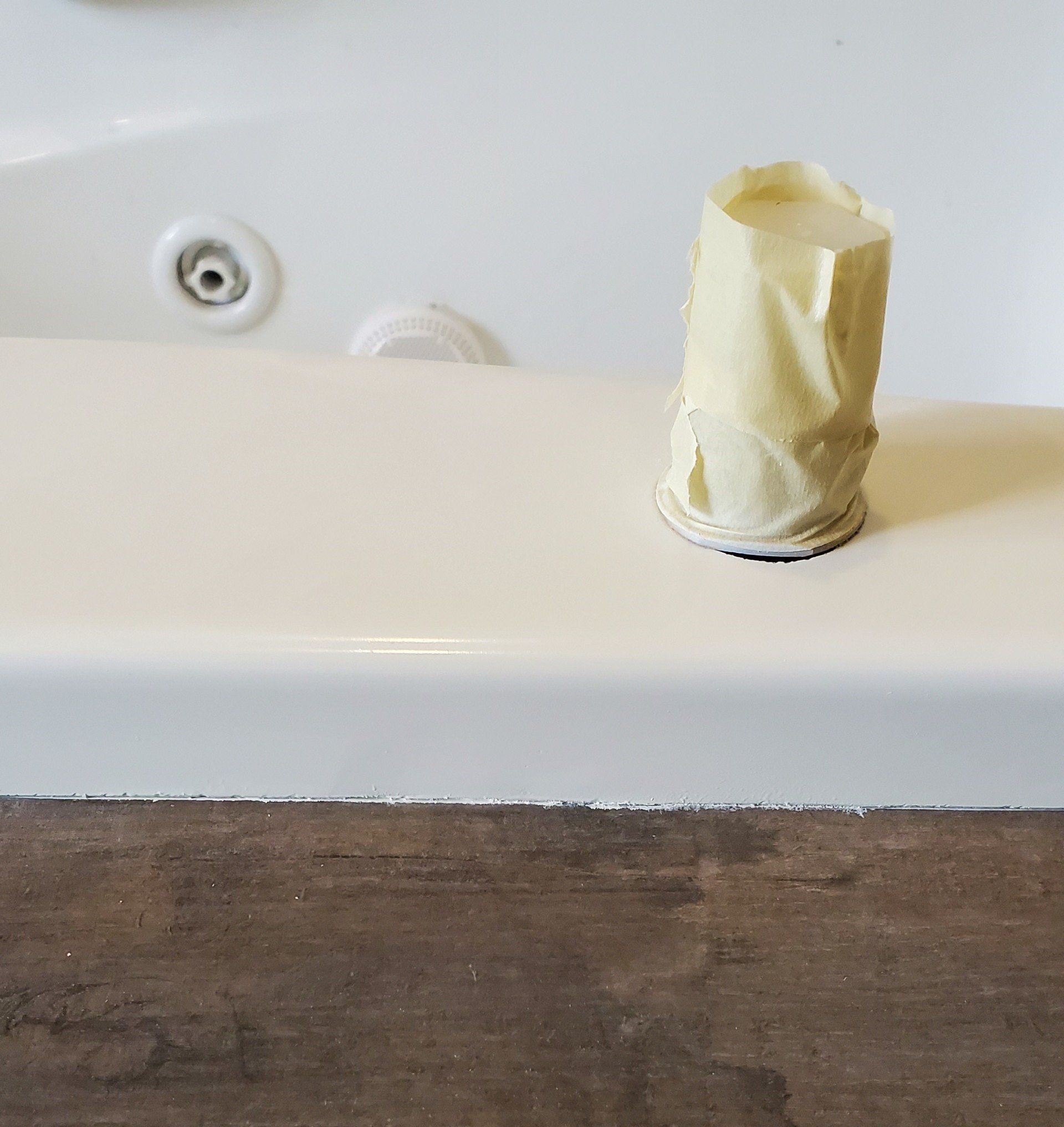 Tub ledge with tape-covered cup, next to a jacuzzi jet. Brown floor beneath the tub.