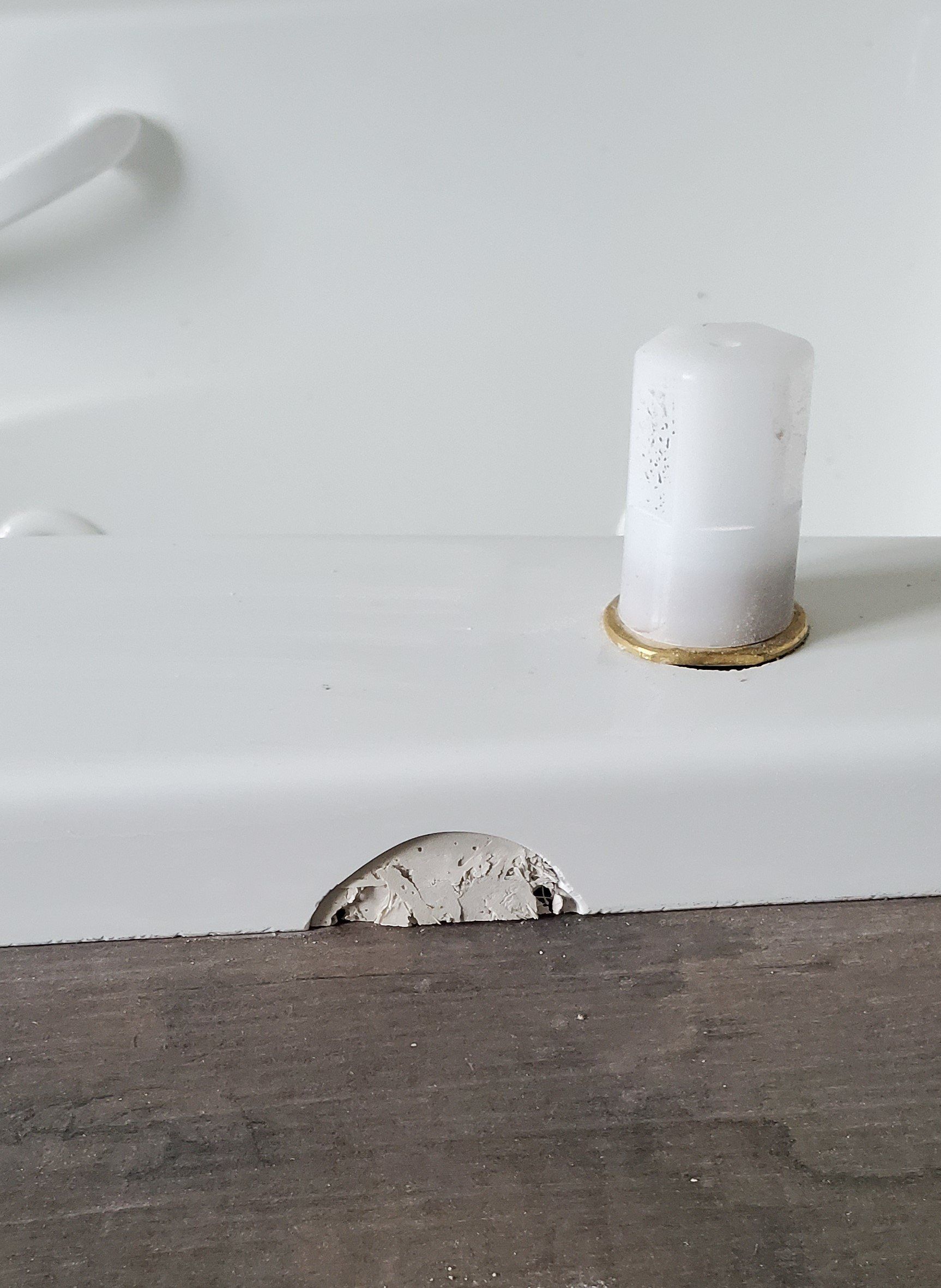 White bathtub rim with a damaged section near the floor. A white candle sits on the rim.