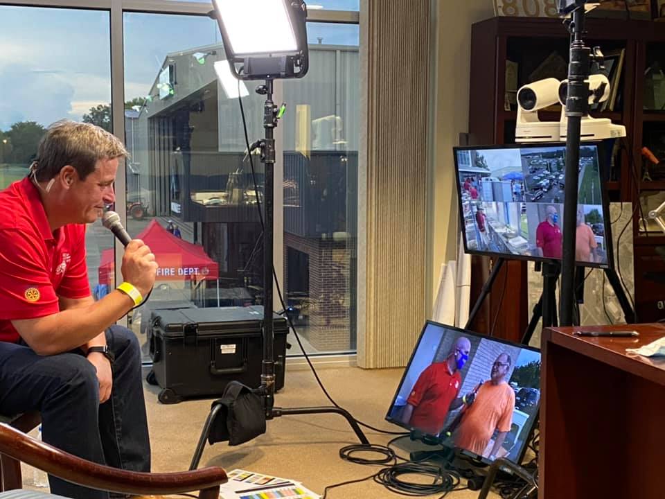 A man in a red shirt is talking into a microphone in front of two monitors.