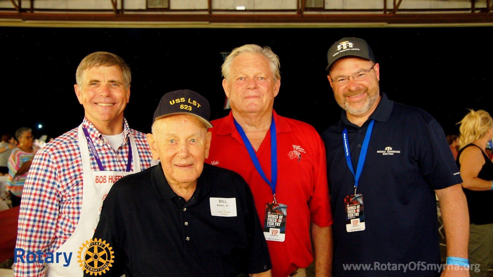 Four men are posing for a picture with one wearing a rotary apron
