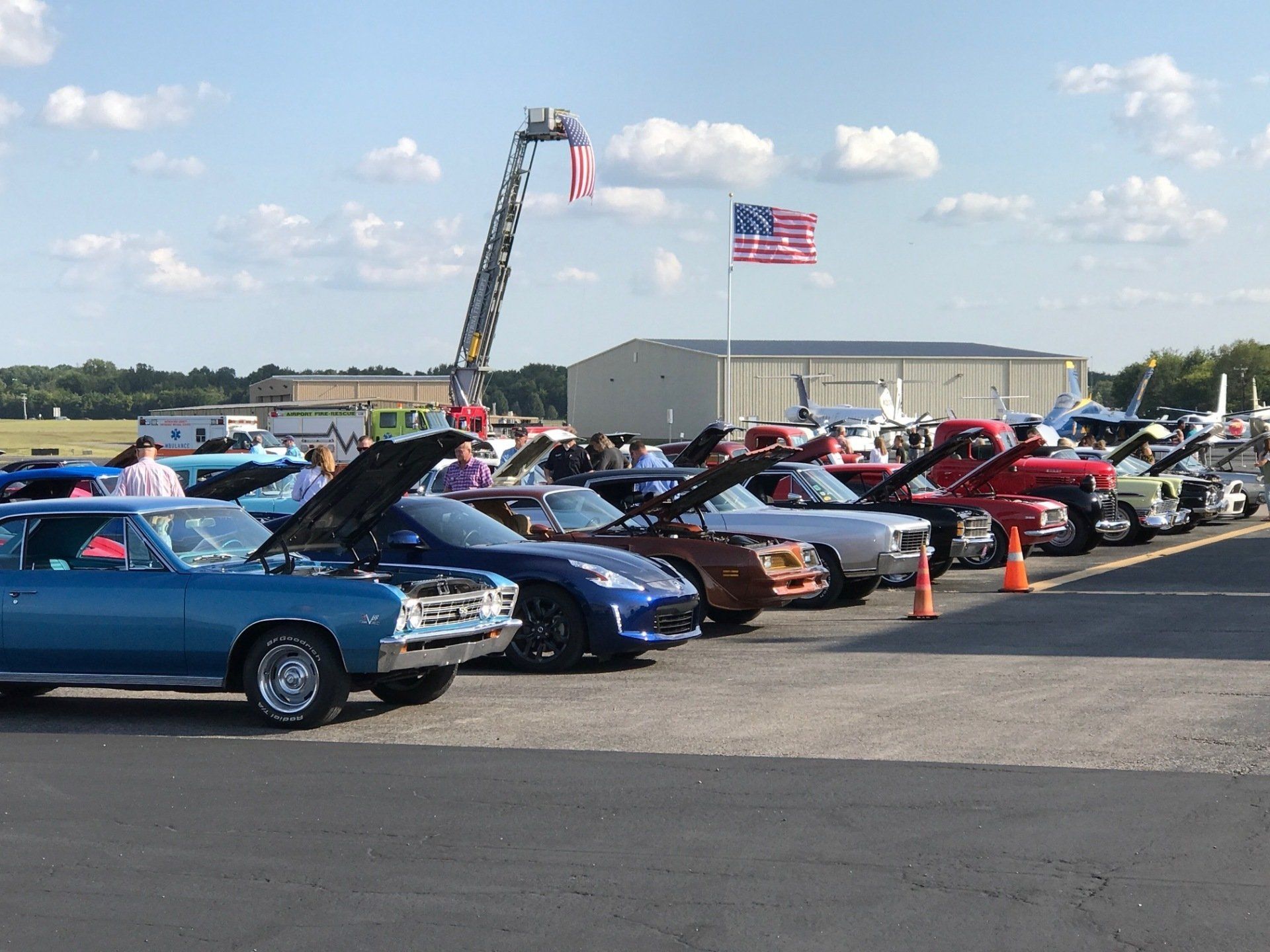 A row of cars are parked in a parking lot at a car show.