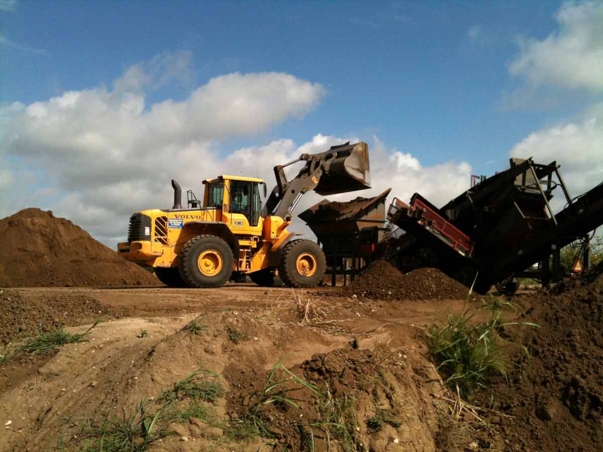 Yellow loader dumping soil into a soil processing machine against a blue sky.
