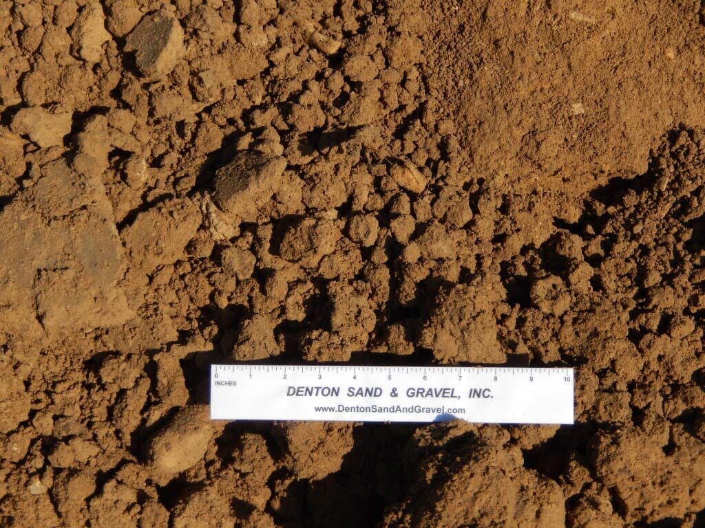 Brown soil sample with visible chunks, a ruler rests on the ground for scale.