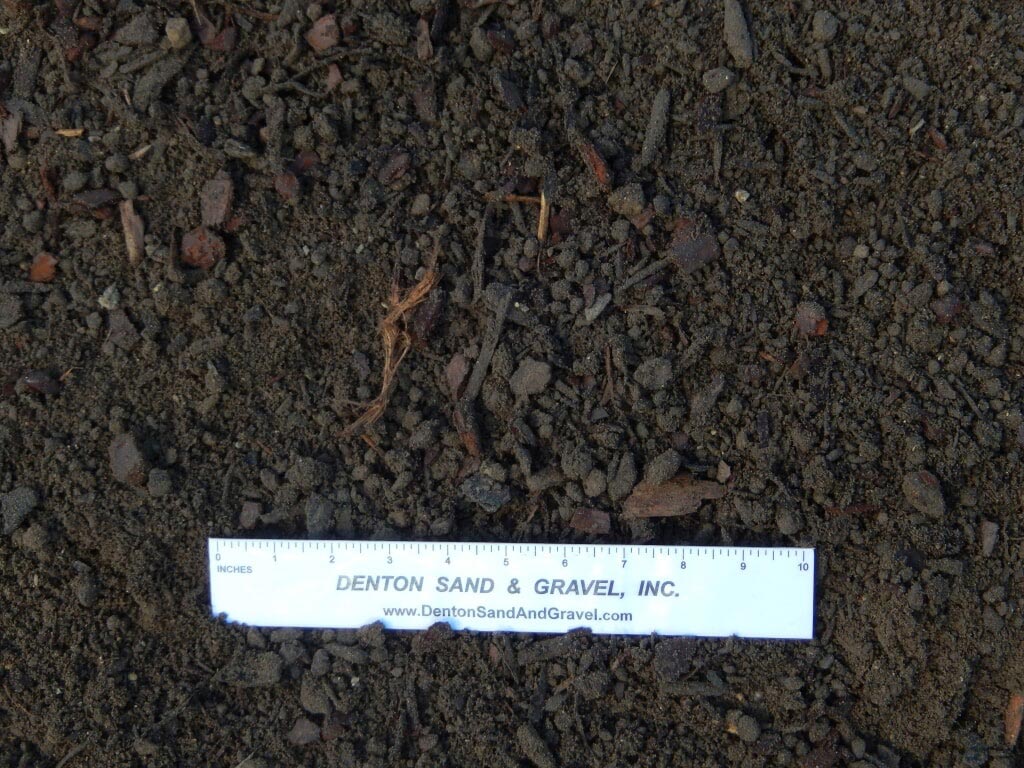 Dark brown soil with small rocks and pieces of wood, a ruler with the company name on it.