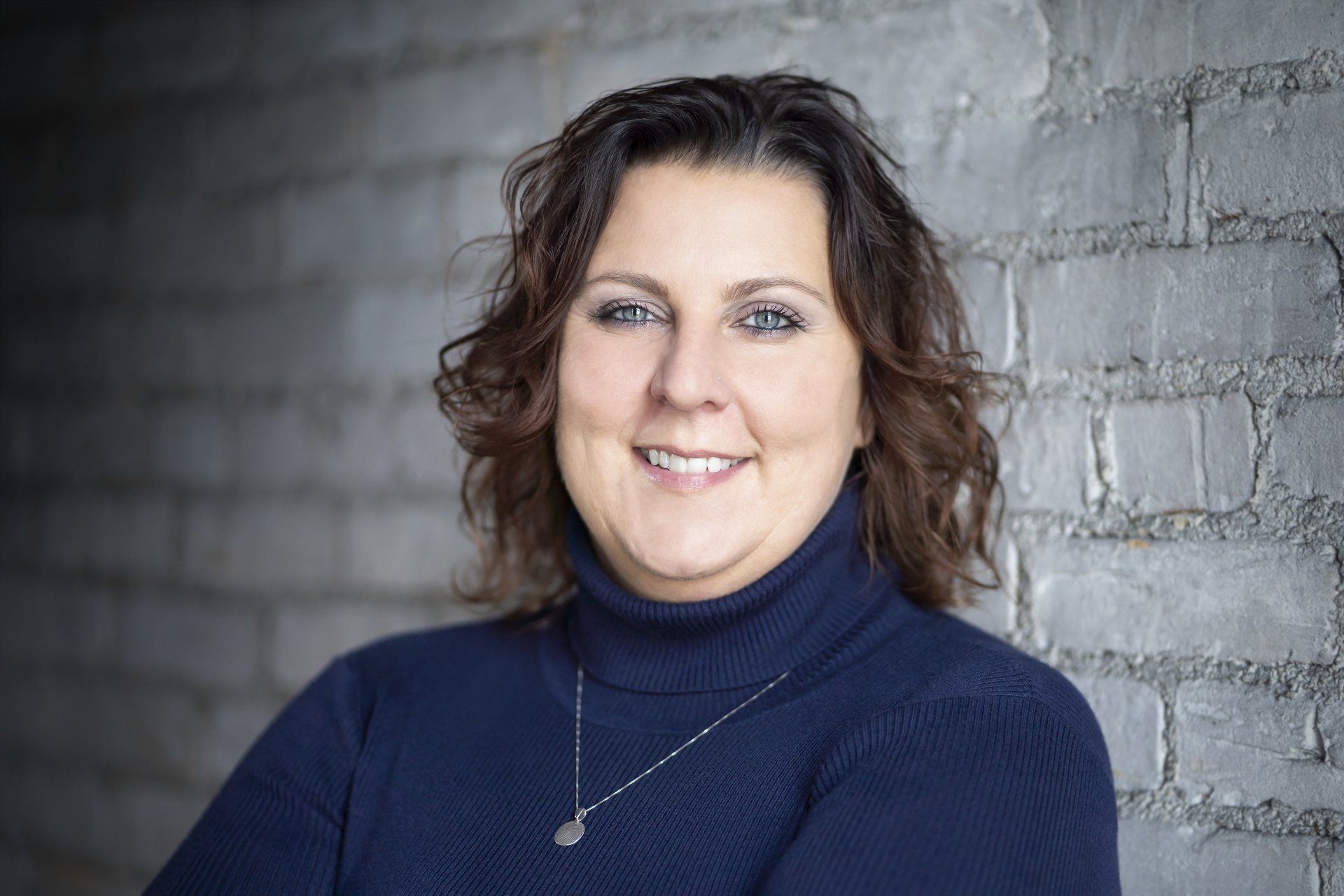 Woman with short brown hair smiles, leaning against a gray brick wall, wearing a blue turtleneck.