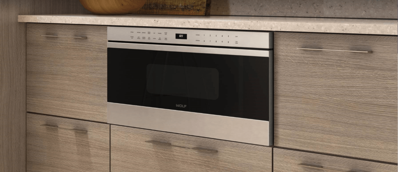 Horno microondas empotrado de acero inoxidable tipo cajón, instalado en un mueble de cocina de madera clara debajo de la encimera.