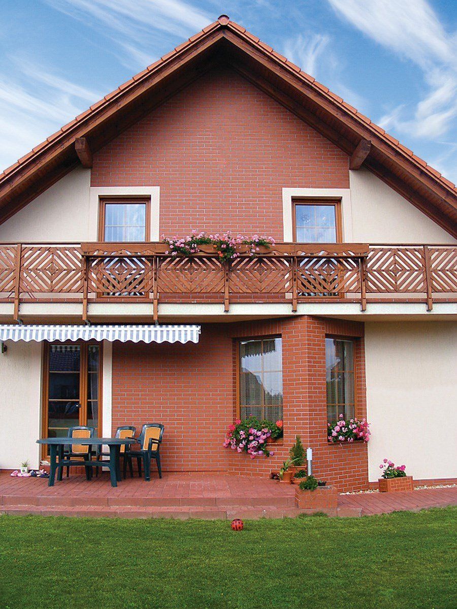 Two-story house with brick facade, balcony with flowers, outdoor dining, and green lawn.