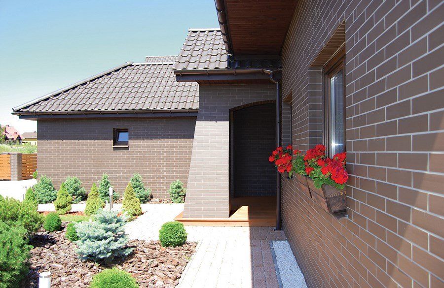 Brown brick house with dark roof, small shrubs and flowers.