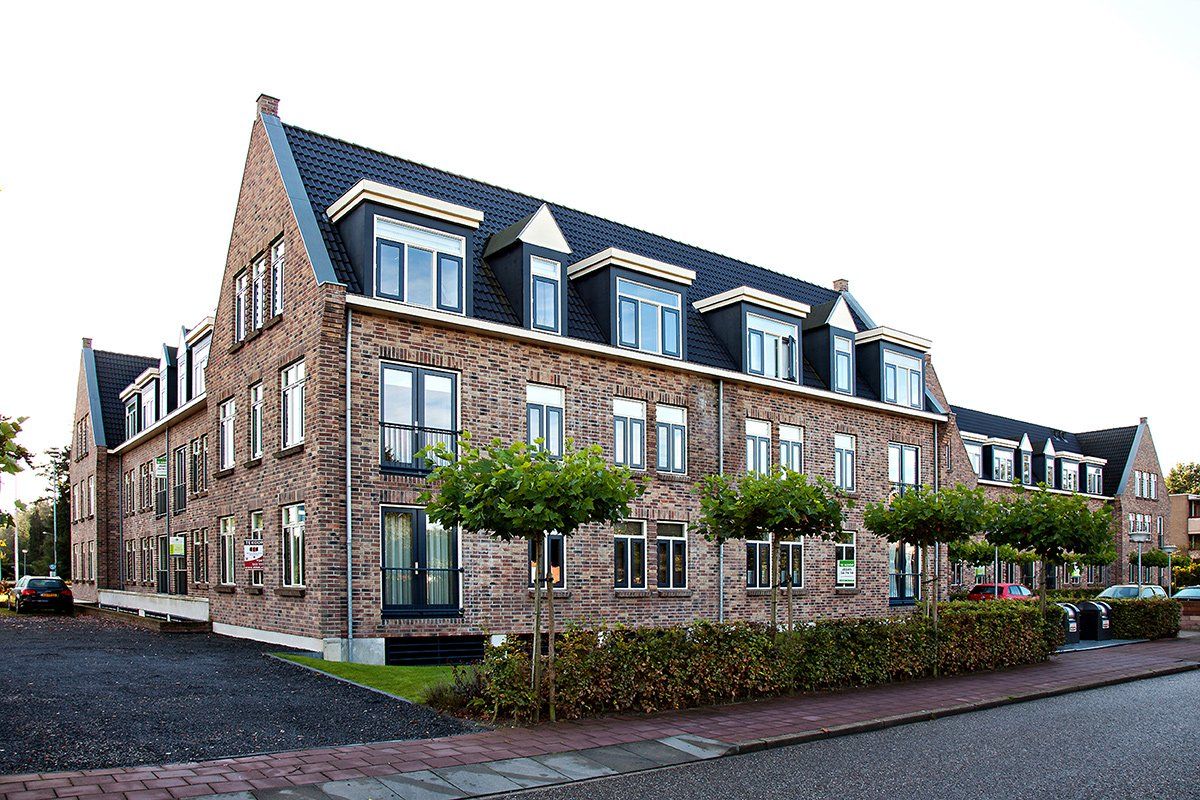 Brick apartment building with dark roof and multiple dormer windows, row of small trees in front.