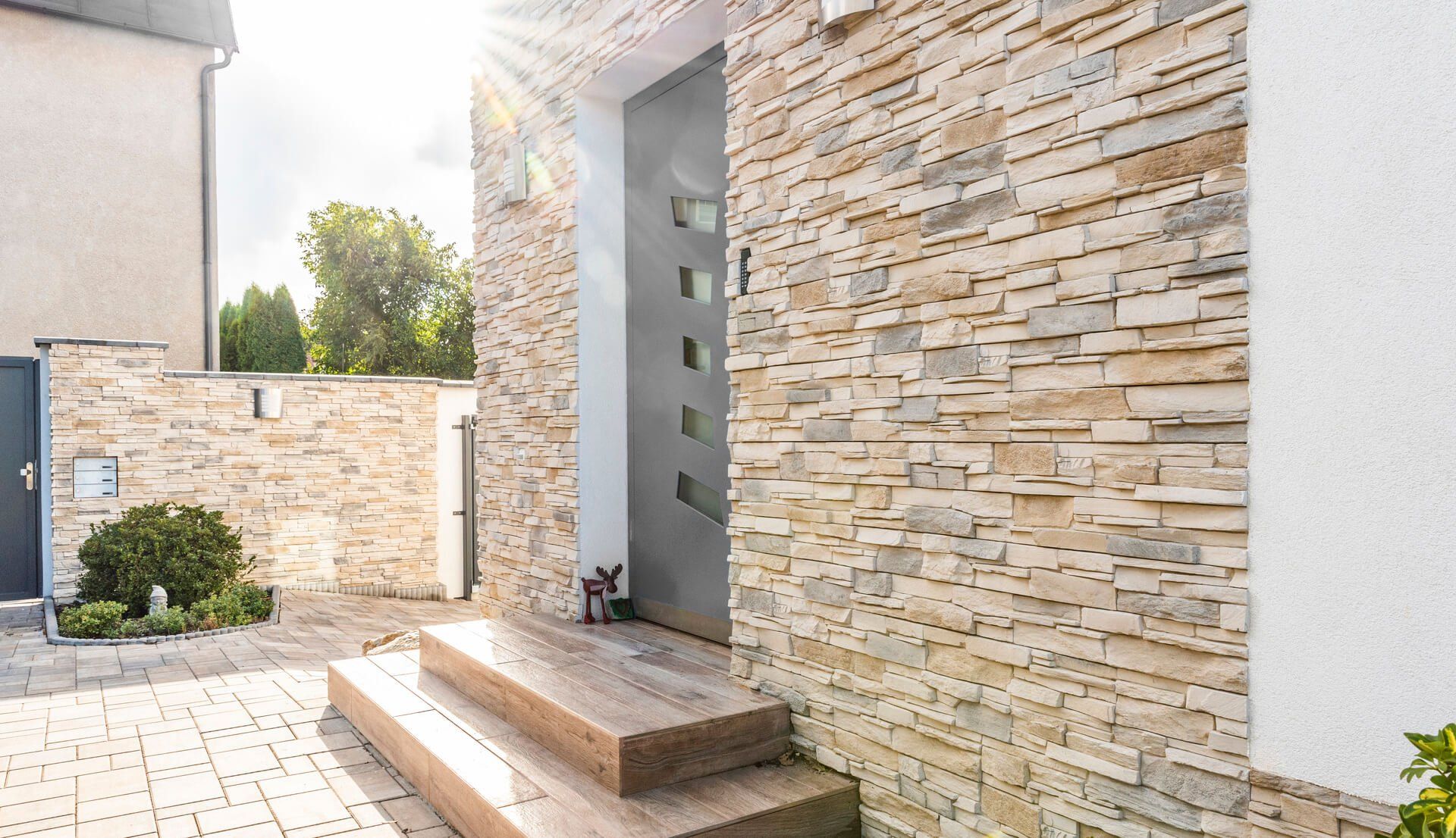 Modern house entrance with stone facade, gray door, wooden steps, and a glimpse of a small dog.