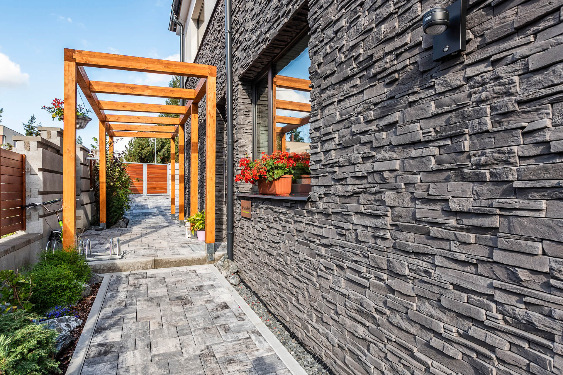 Dark gray brick wall with wooden pergola over a stone walkway.