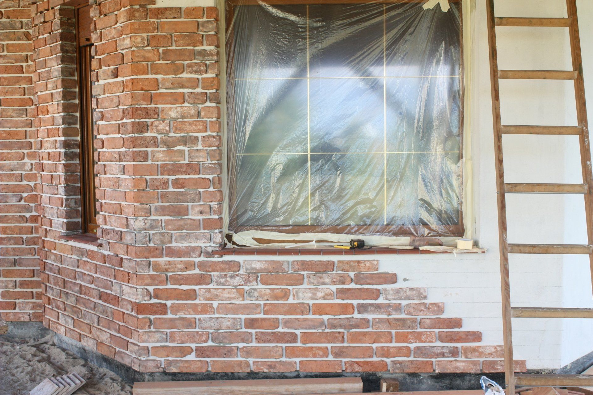 Brick exterior corner of a building under renovation, window covered in plastic, wooden ladder.