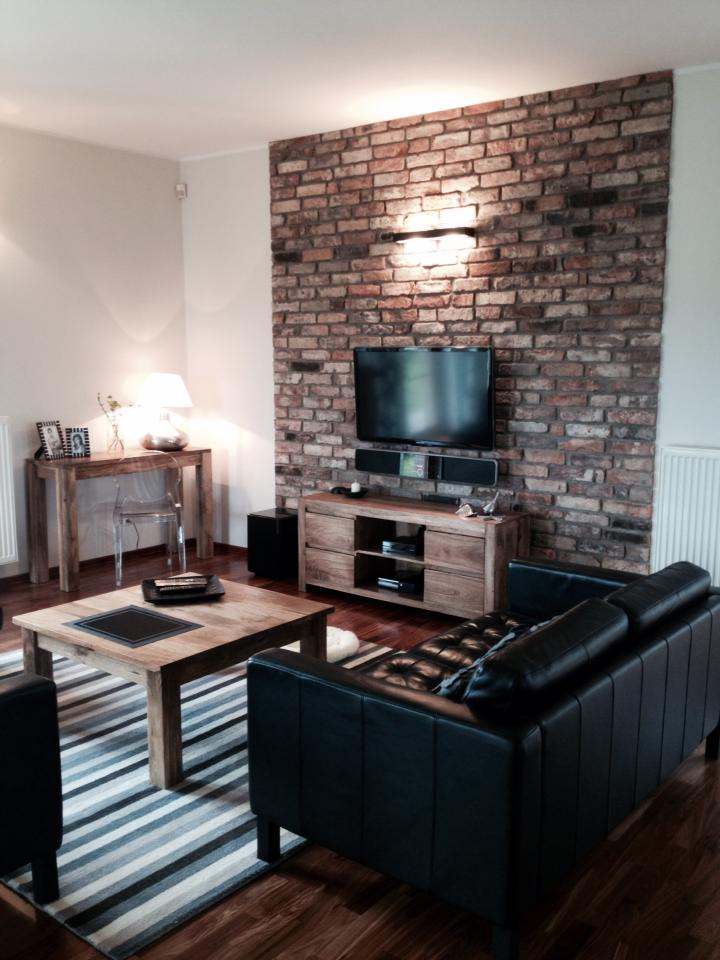 Living room with brick accent wall, TV, wooden furniture, black leather couch, striped rug.