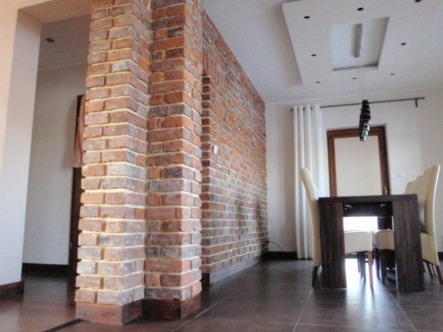 Interior view: Brick wall separates a dining area from a hallway. Dark table and chairs present. White walls, ceiling lights.
