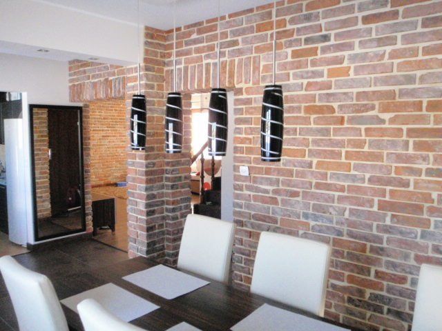 Dining room with brick walls, black and white pendant lights, and white chairs around a dark table.