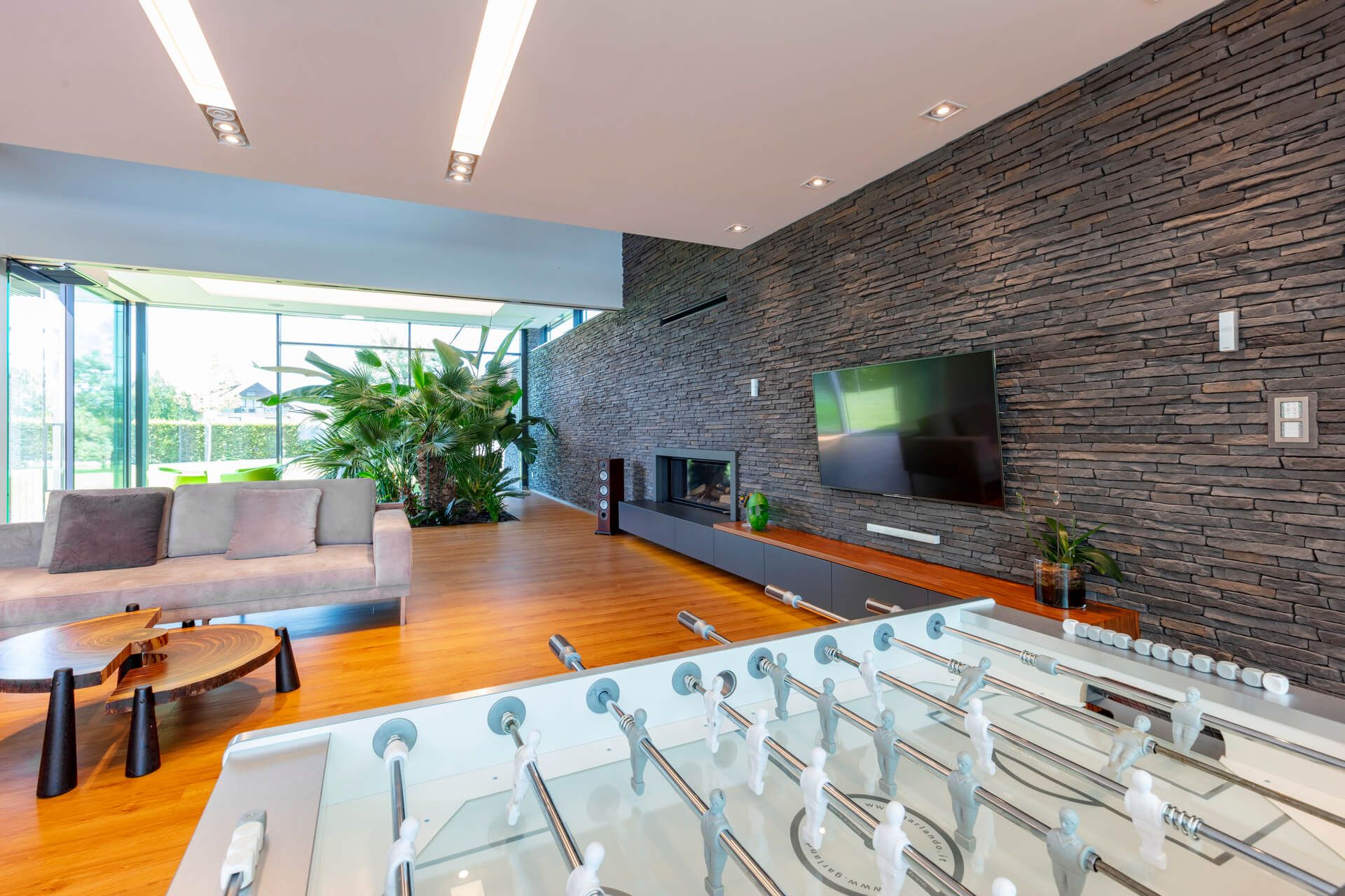 Modern living room with foosball table, stone accent wall, large TV, and view of greenery.