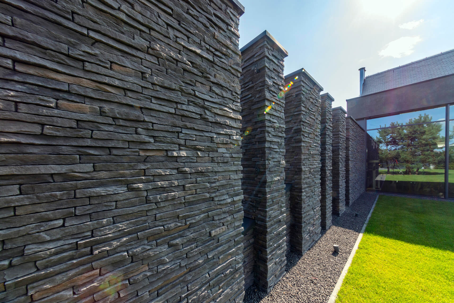 Stone wall with rectangular pillars, constructed of dark gray, layered stone in a sunny outdoor setting.
