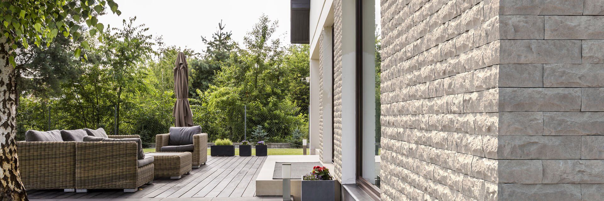 Patio with woven furniture and brick wall, lush greenery in the background.