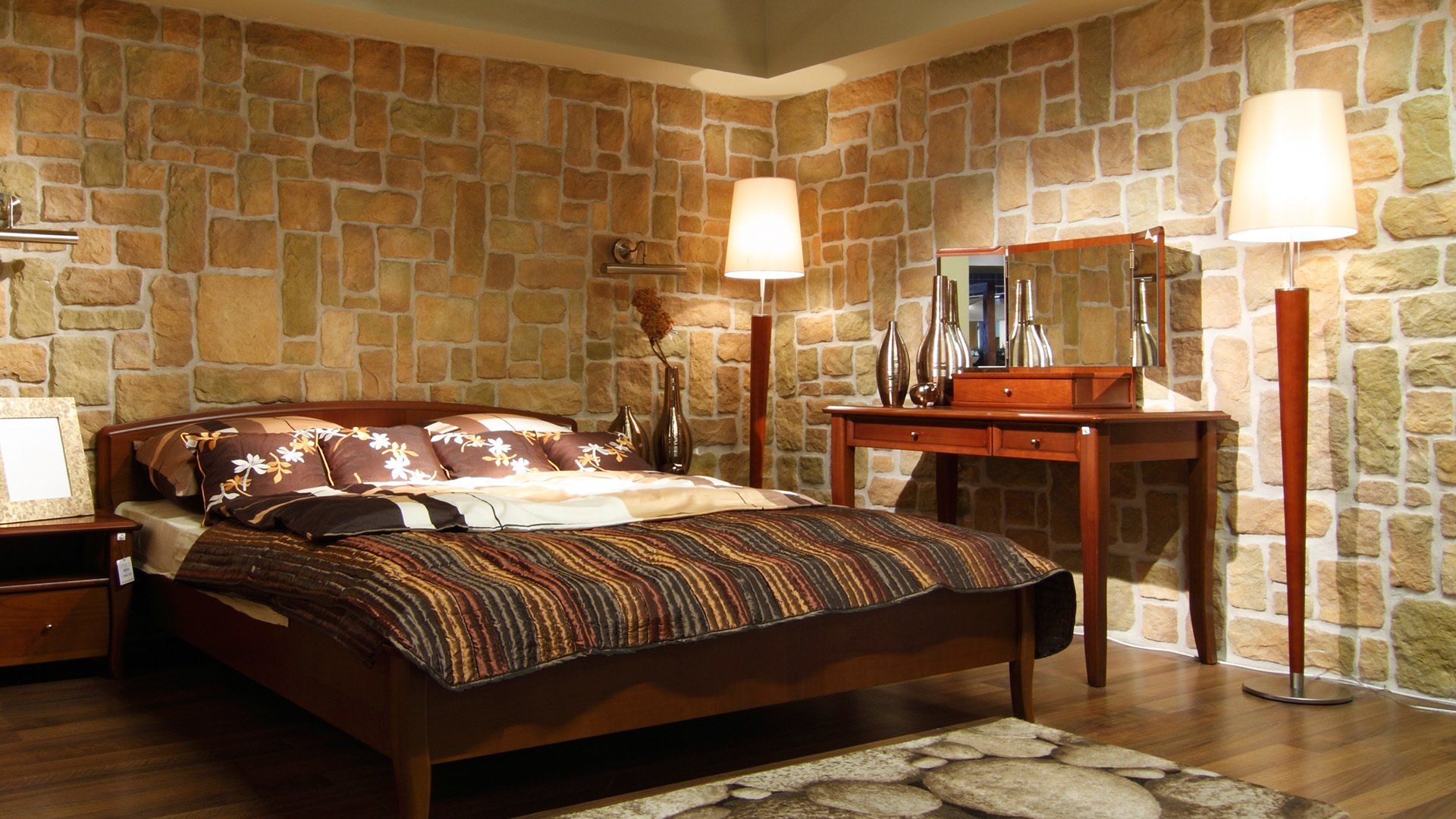 Bedroom with a wooden bed, nightstand, and dresser against a stone-like wall, lit by two lamps.