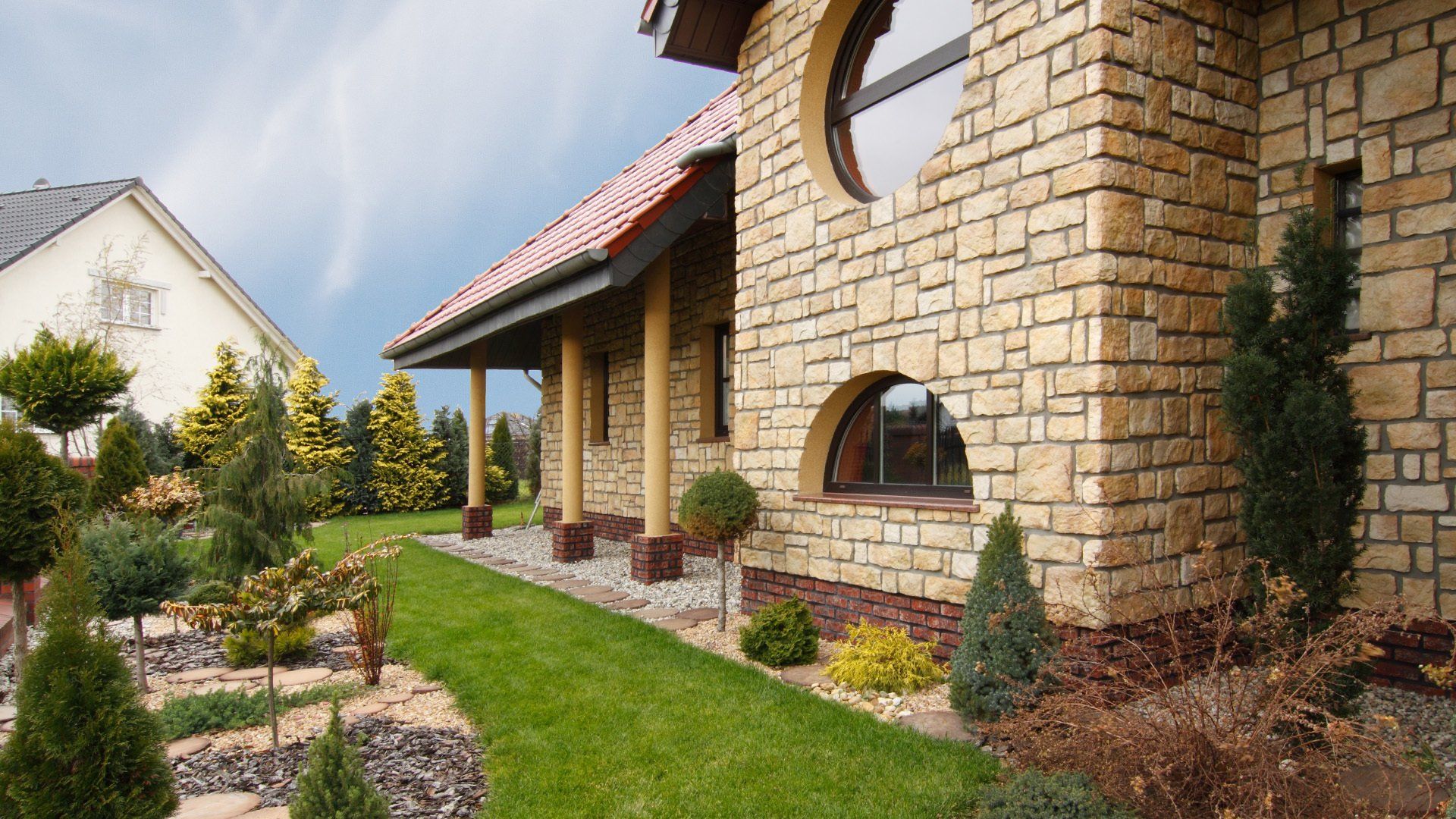 Stone-faced house with porch, green lawn, trees, and sky.
