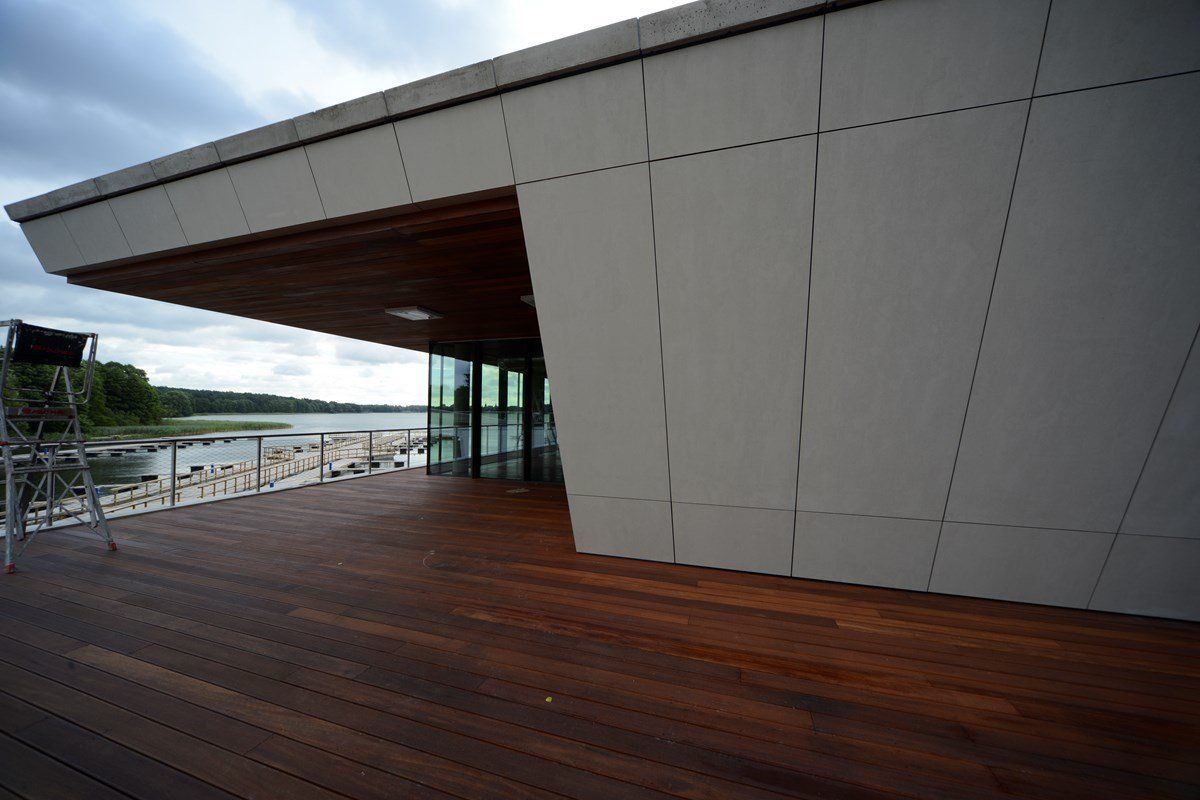 Modern building with wooden deck and large glass windows overlooking water. Gray walls and overcast sky.