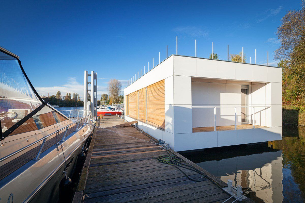 White floating rectangular building with wooden shutters, docked near a boat on a river, under a blue sky.