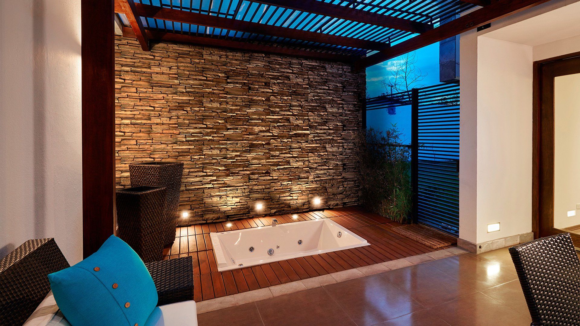 Outdoor spa area with wooden deck, stone wall, and jacuzzi. Lit by recessed lights.