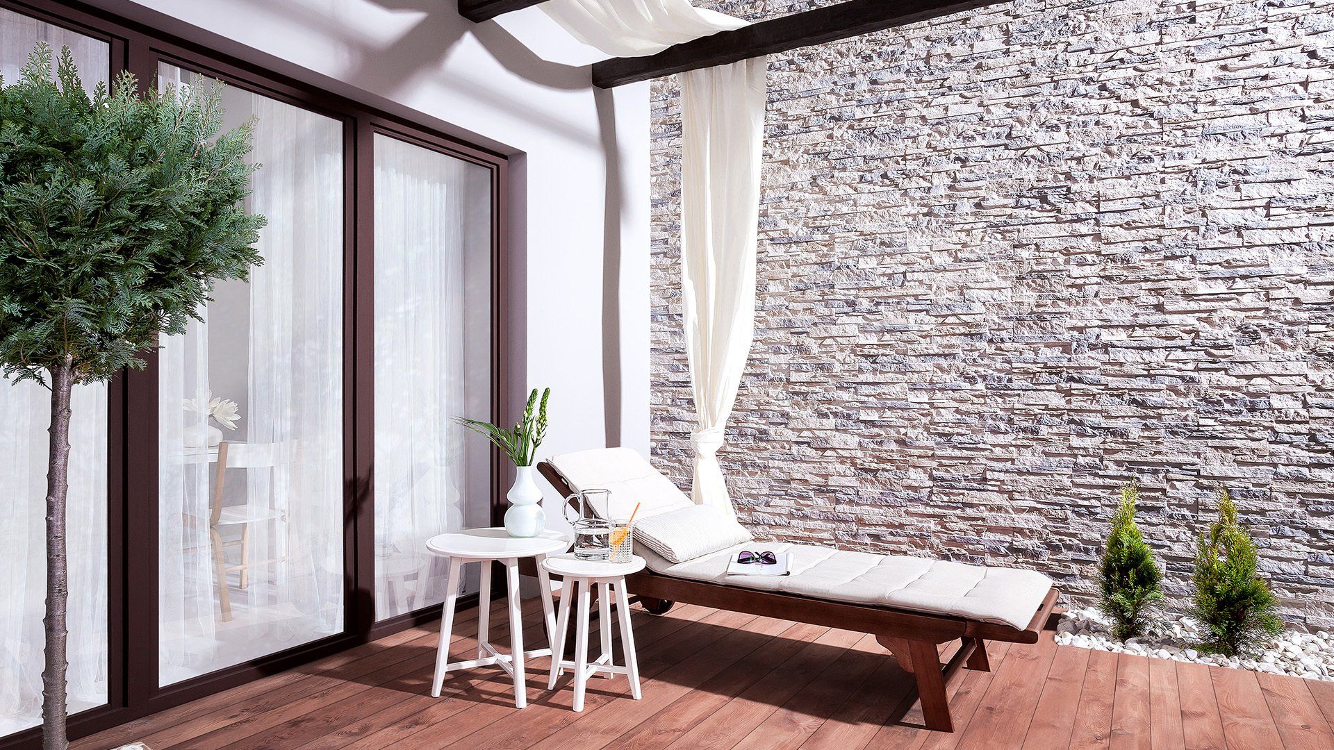 A relaxing outdoor patio with a chaise lounge, white curtains, and a stone wall.