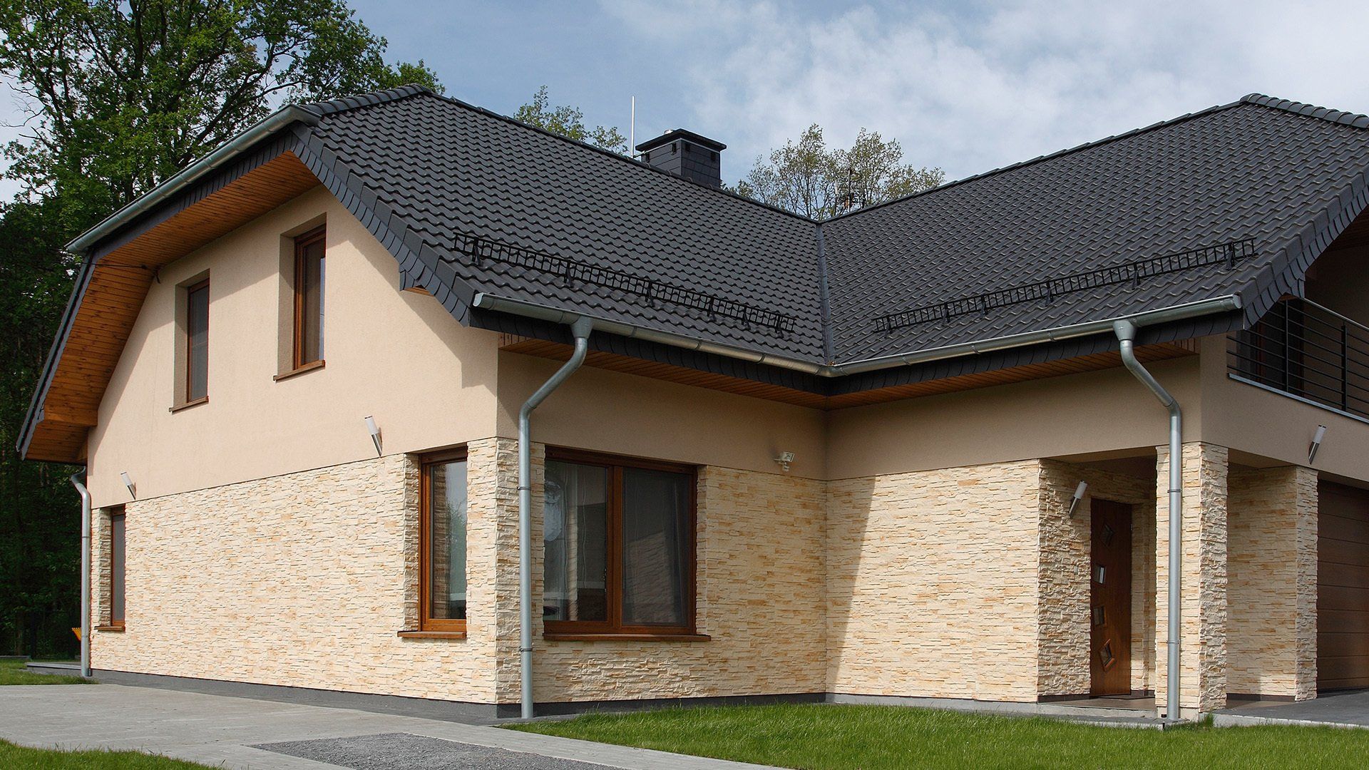 Beige house with a black roof and stone facade. Set in green grass with trees.