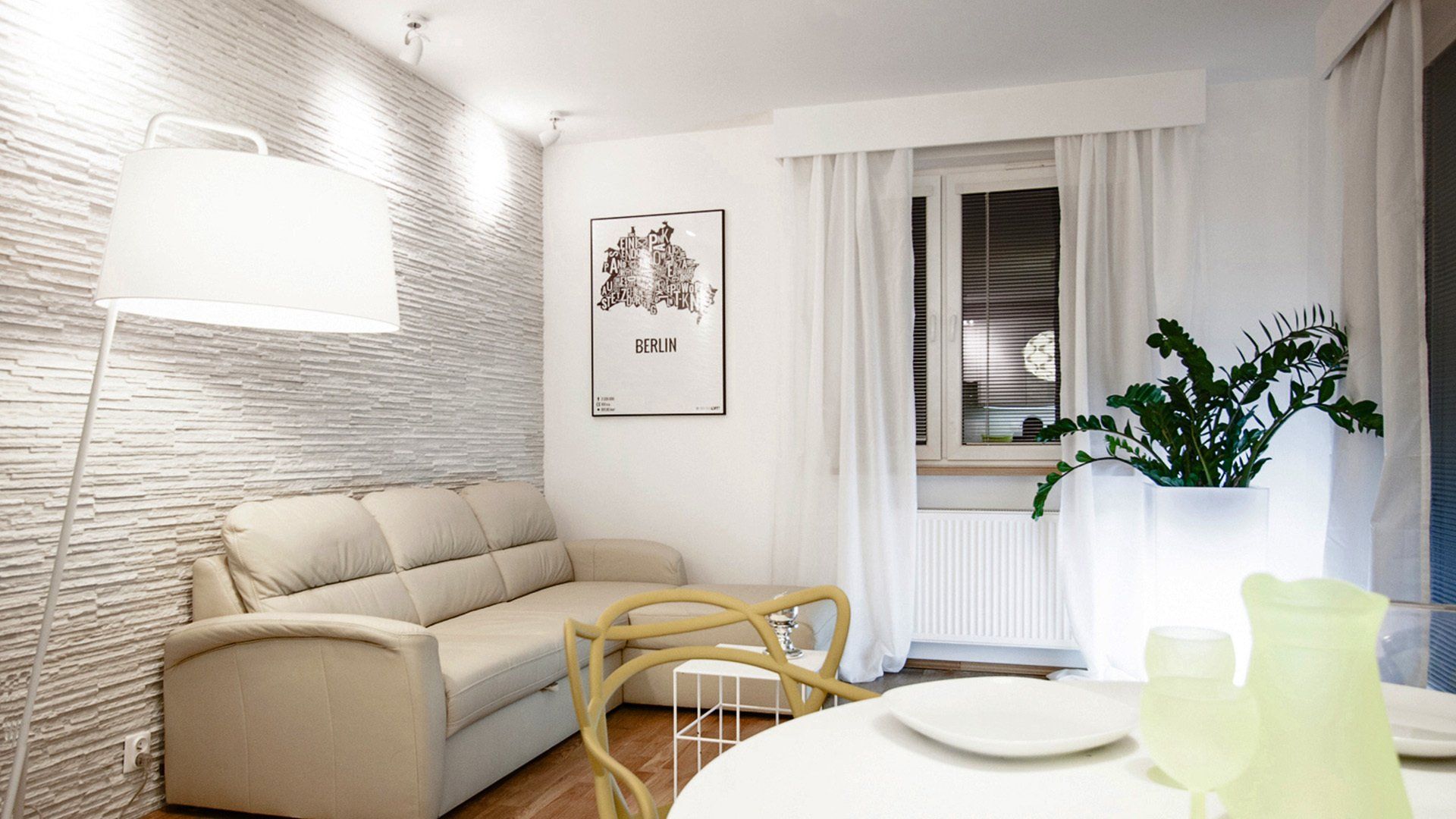 Cozy living room with beige sofa, textured wall, large lamp, and a dining table by a window with white curtains.