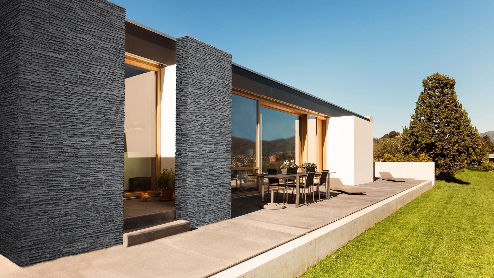 Modern home exterior with dark stone accents, large windows, and a patio with outdoor dining set.