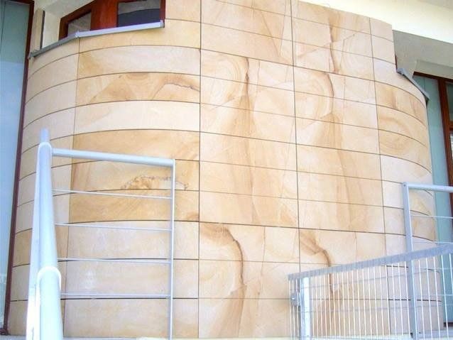 Sandstone-tiled curved wall with horizontal and vertical lines, metal railing, and windows.